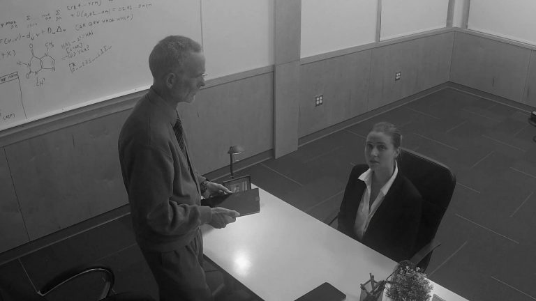 Still from indie short film Cynthetic. Actor Jenna Hill's character looks up towards the CCTV camera feed that the audience is viewing an interview through, in black-and-white.